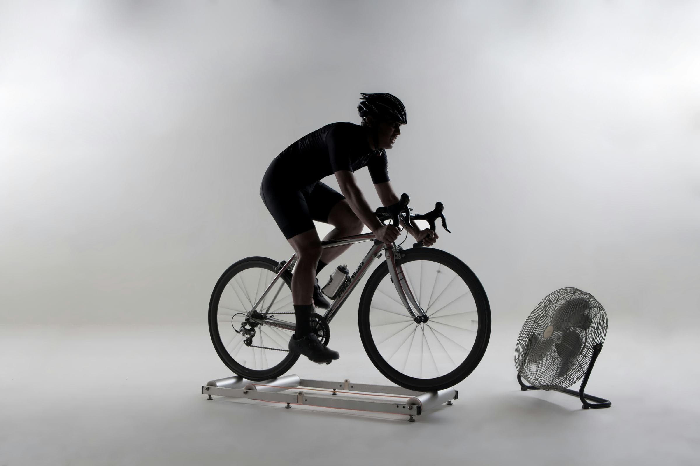 Ciclista pedaleando sobre rodillos en un estudio, con un ventilador delante.